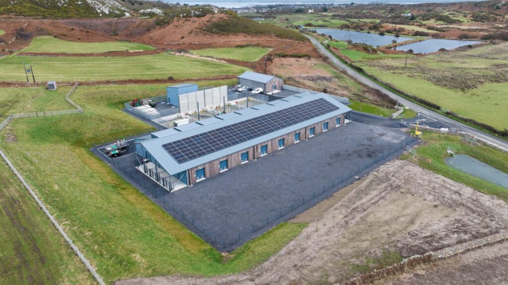 A large building with solar panels on the roof, surrounded by green fields.