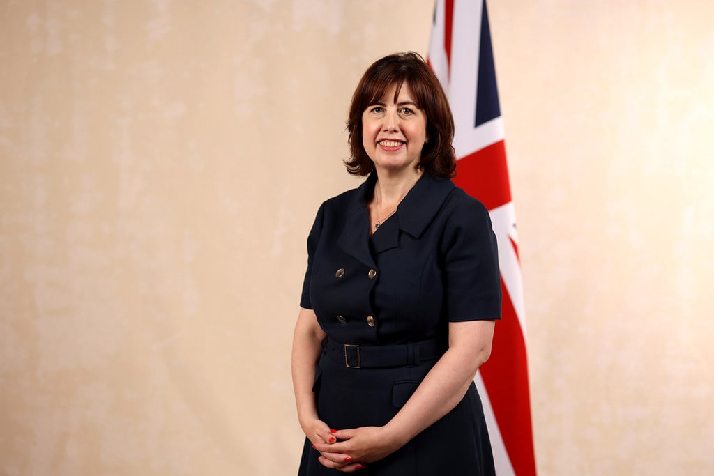 A white woman in a short sleeved black dress stands in front of a Union Flag