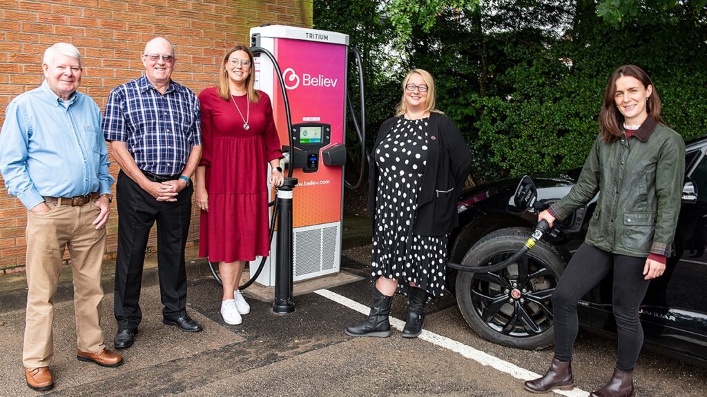 A group of white men and women stand in front of a Believ branded EV charger