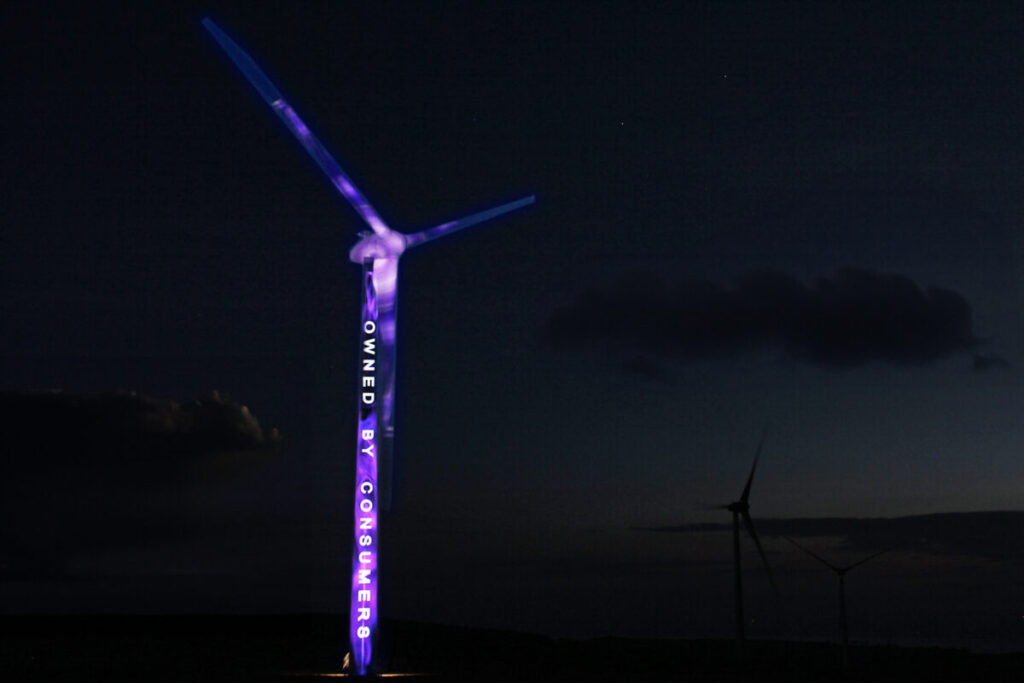 A wind turbine at night, with purple light and text reading "owned by consumers" projected onto it