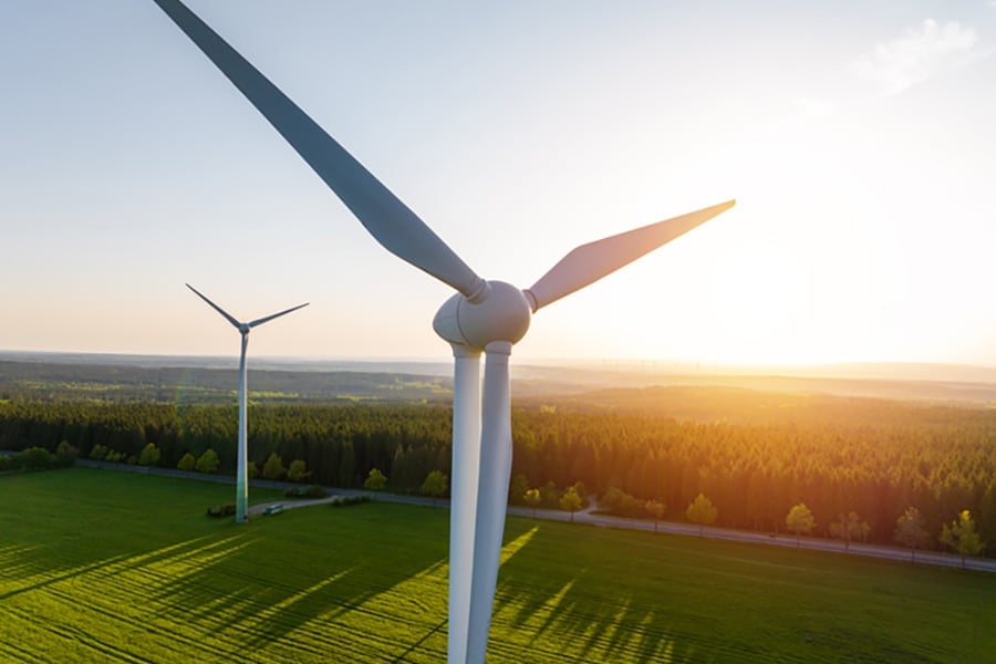 Two wind turbines, one close up and one in the distance, stood on green grass while a setting sun casts long shadows