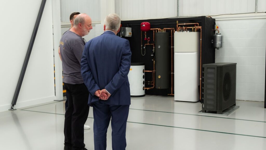 Three men stand with backs to camera looking at a demonstration heat pump model