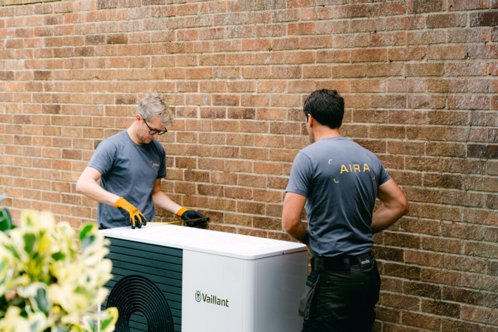 Two men install an air source heat pump next to a brick wall