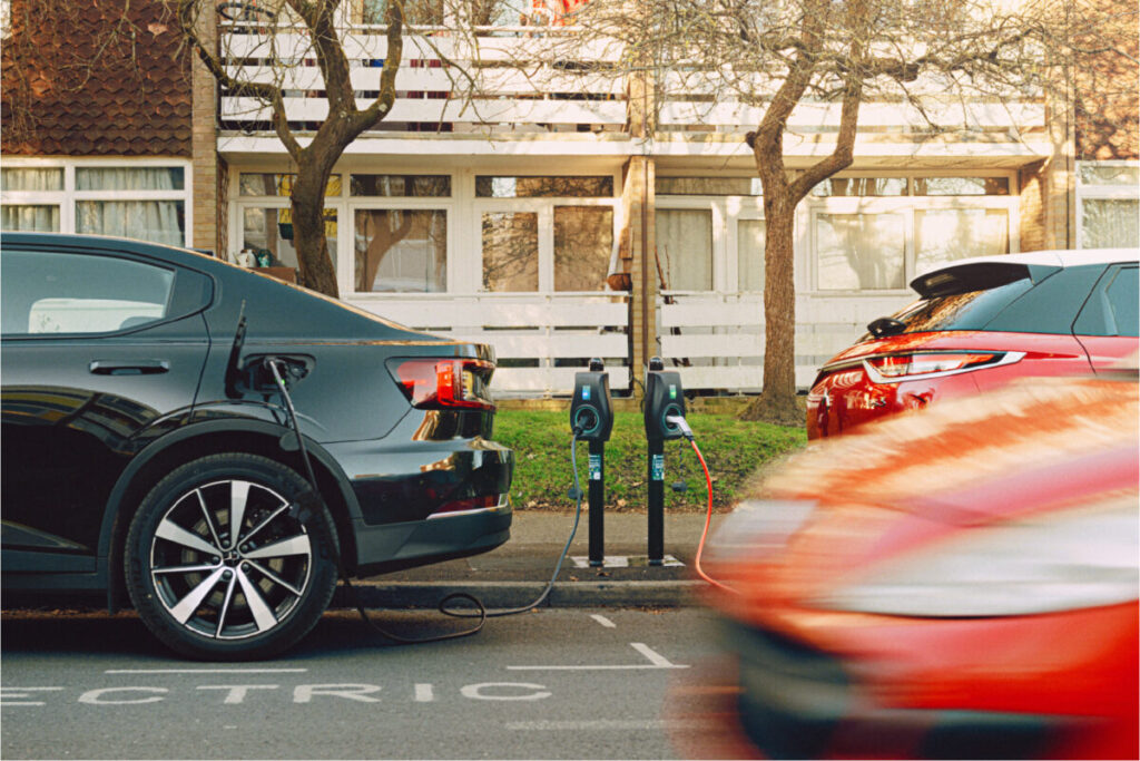 Two cars plugged into an EV chargepoint as one, blurred from speed, drives past