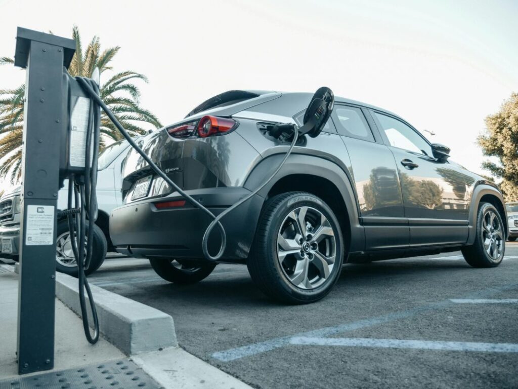 A grey car plugged into an outdoor EV charger with palm trees behind