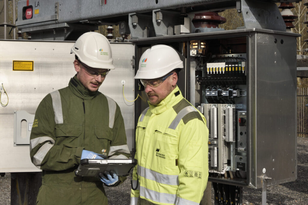 Two men in high vis jackets stand by a large fuse box