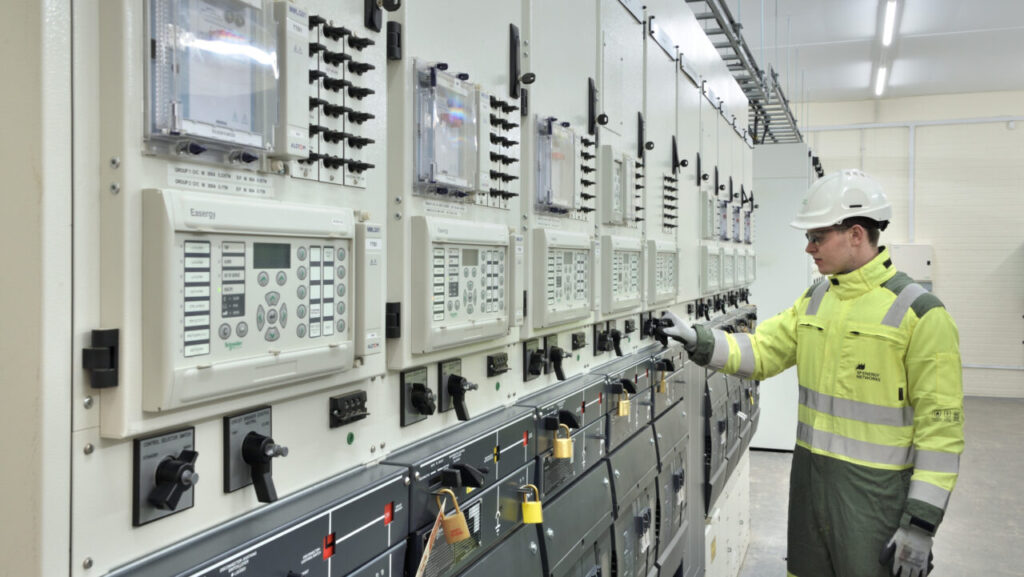a man in a helmet and hi vis vet looks at a wall of switches