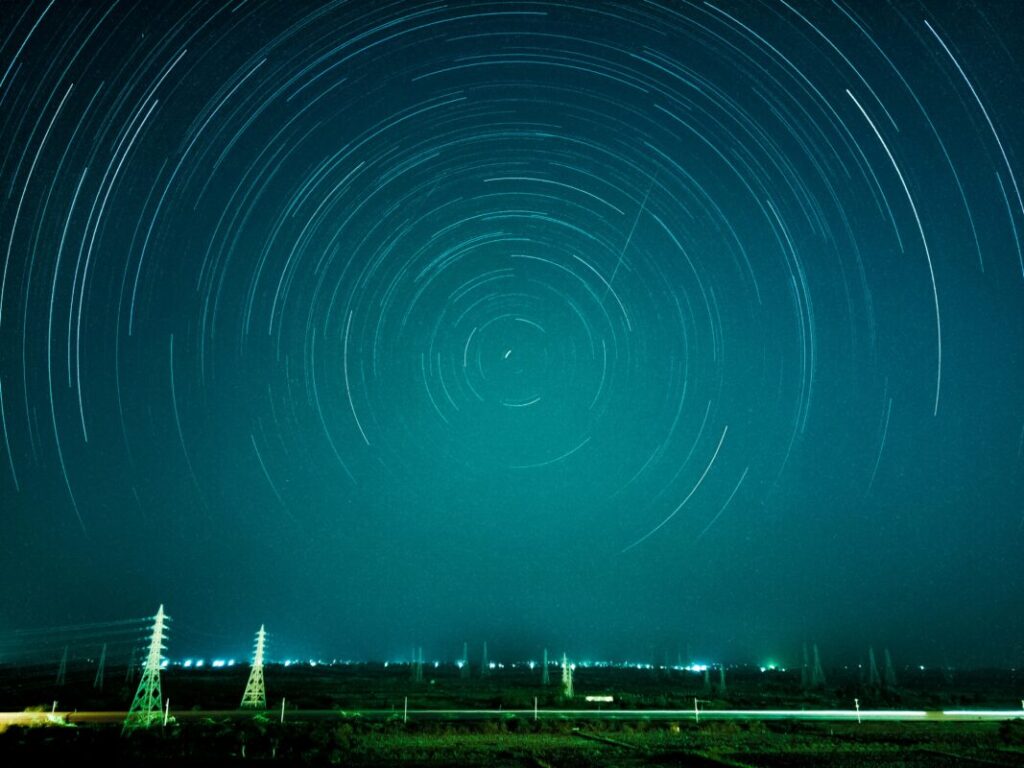 Several pylons in the distance in front of a green blue sky with trails of light in circles across it