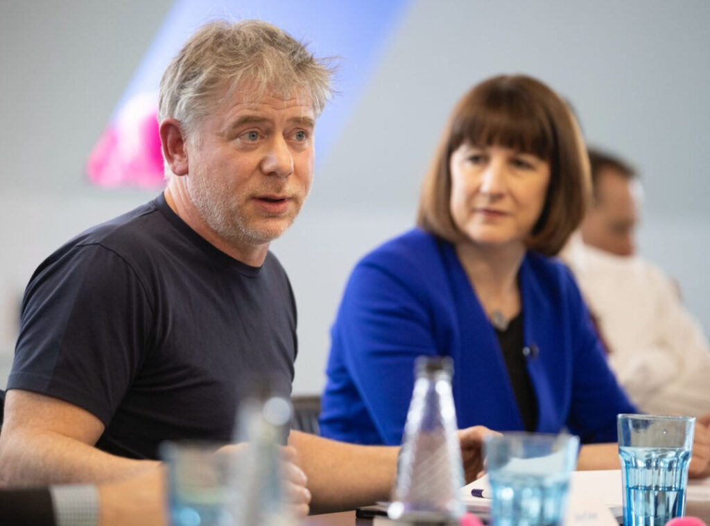 a white middle aged man in a black tshirt sits centre frame, with a woman out of focus behind him