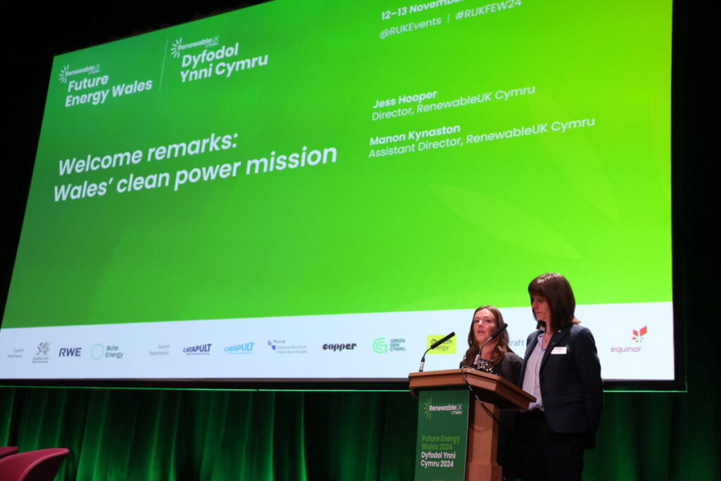 Two women stand in front of a large screen as they present a report at a conference