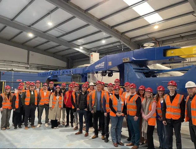 A group of people in hard hats and hi vis vests stand near industrial equipment