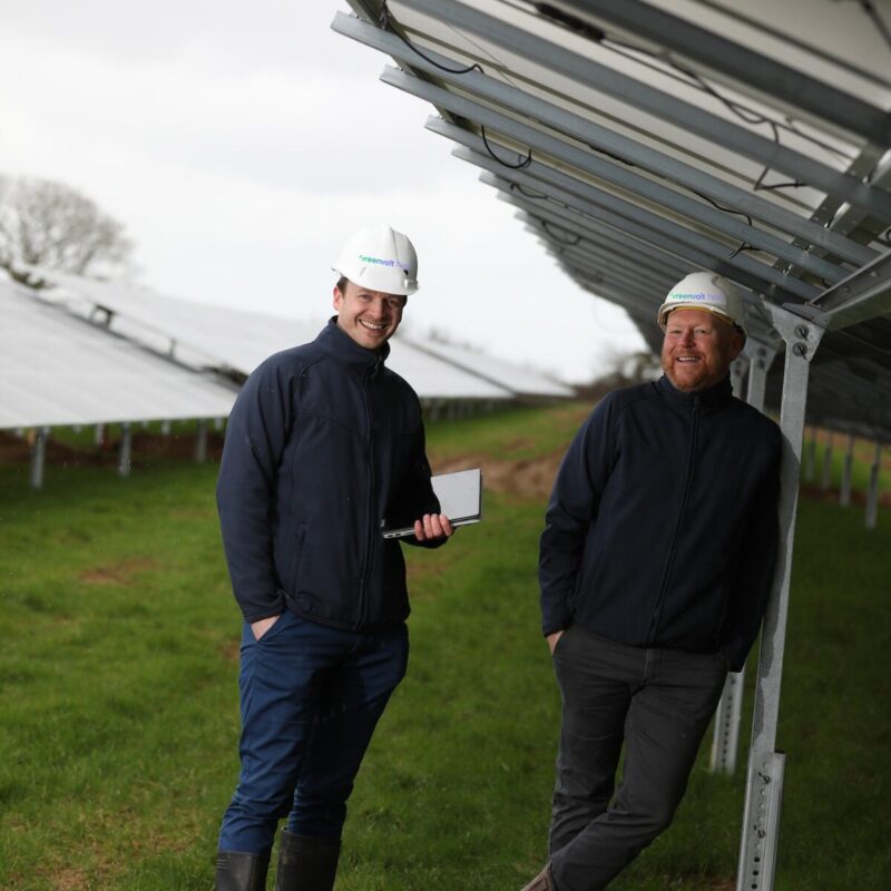 two white men in white helmets smile as they stand under a solar panel