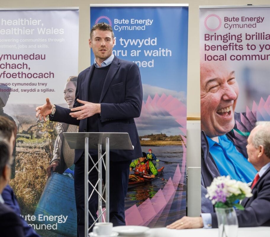 a white man in a suit stands at a lectern in front of advert banners making a speech