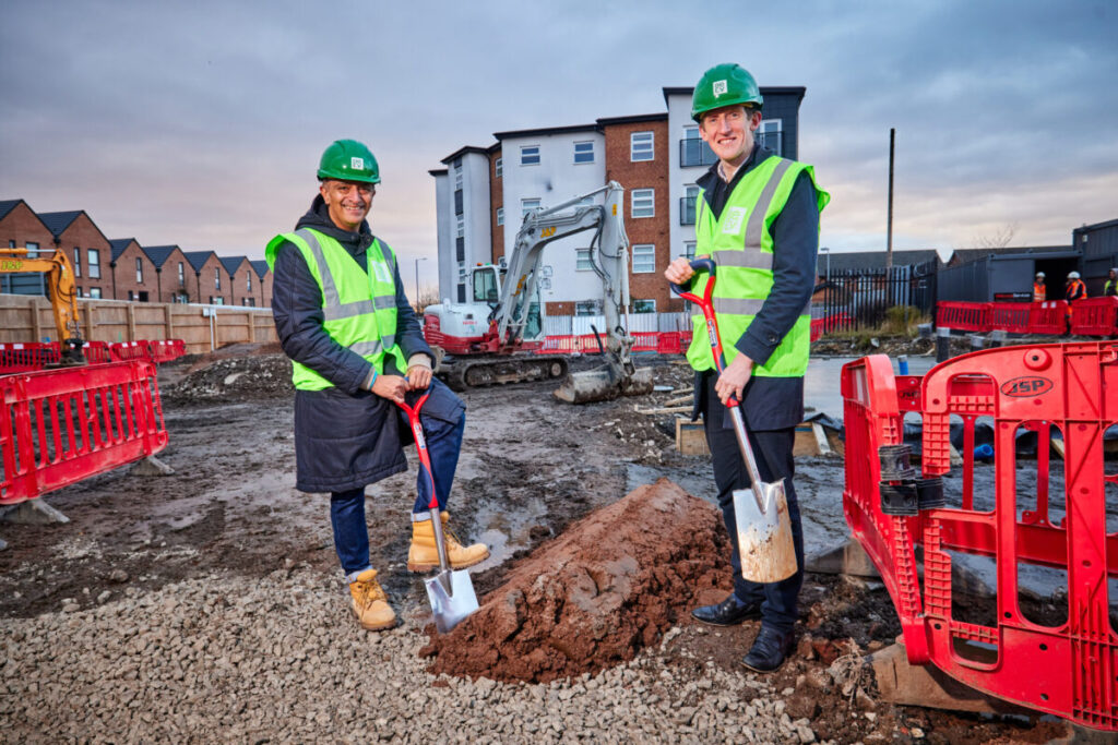 two men in high vis vetss and helmets, one white, one west Asian, hold shovels on a construction site