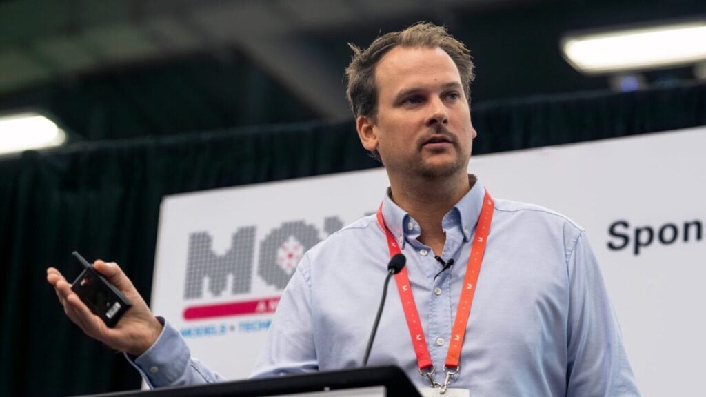 A white man wearing a lanyard stands behind a podium