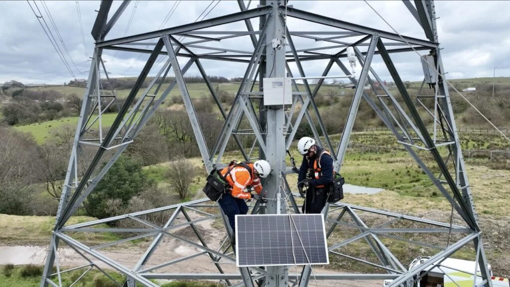 workers install a sensor with a solar panel on a transmission tower