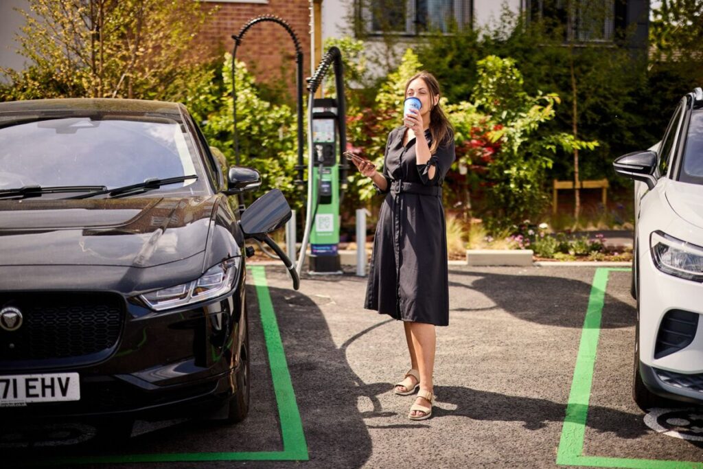 a a white woman in a black dress drinks a takeaway coffee while charging her EV