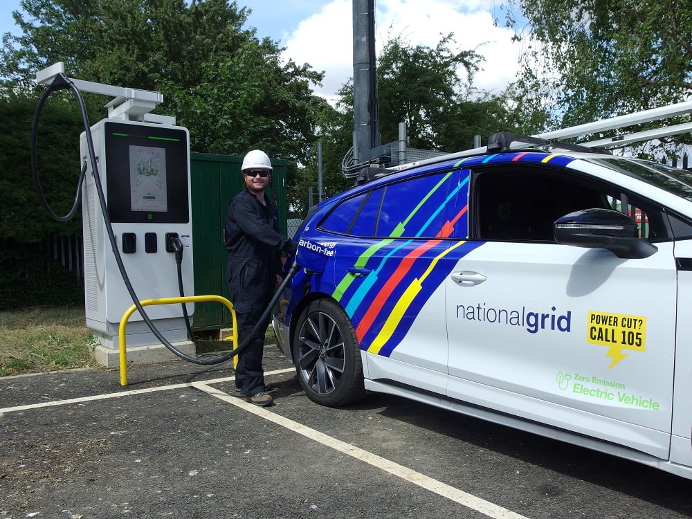 a man in black overalls and a white helmet plugs an EV charger into a National Grid branded EV
