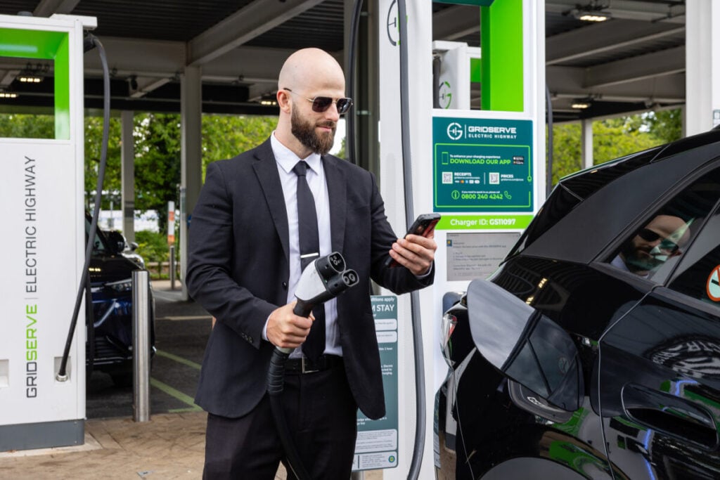 a bald man in sunglasses and a suit checks his phone as he holds an ev charger, ready to plug into his car.