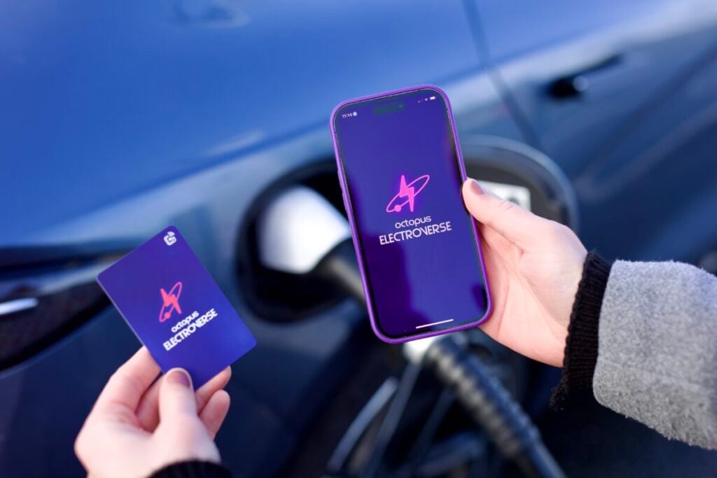 a hand holds a smartphone and a small card, both bearing the Octopus Electroverse logo, with an electric car plugged in to charge in the background