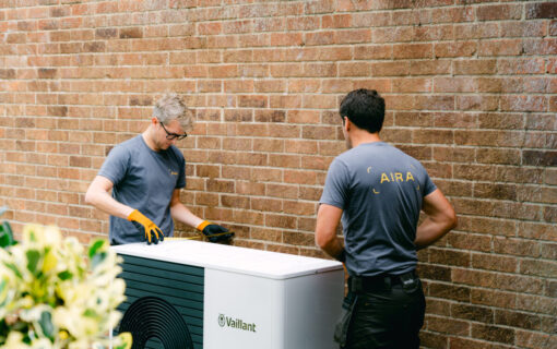 Two men install an air source heat pump next to a brick wall
