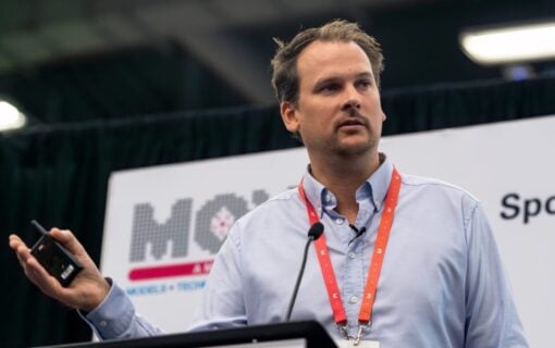 A white man wearing a lanyard stands behind a podium