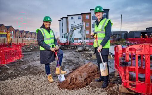 two men in high vis vetss and helmets, one white, one west Asian, hold shovels on a construction site