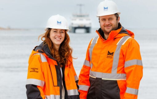 a white woman and a white man wear orange high vis jackets and white helmets as they stand by the sea