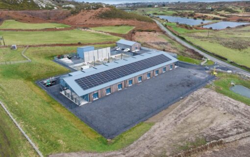 A large building with solar panels on the roof, surrounded by green fields.