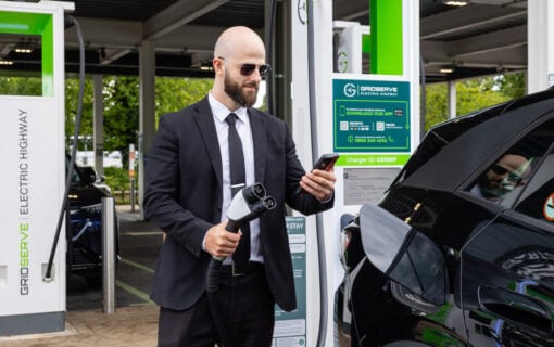 a bald man in sunglasses and a suit checks his phone as he holds an ev charger, ready to plug into his car.