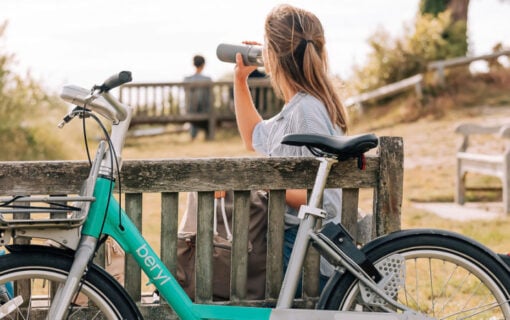 A blue ebike leans against a wooden bench where a woman sits and sips water