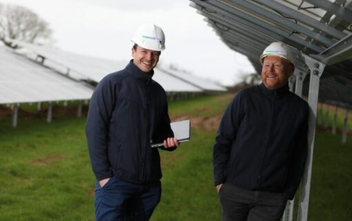 two white men in white helmets smile as they stand under a solar panel