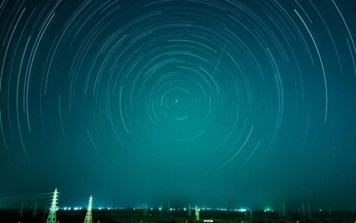 Several pylons in the distance in front of a green blue sky with trails of light in circles across it