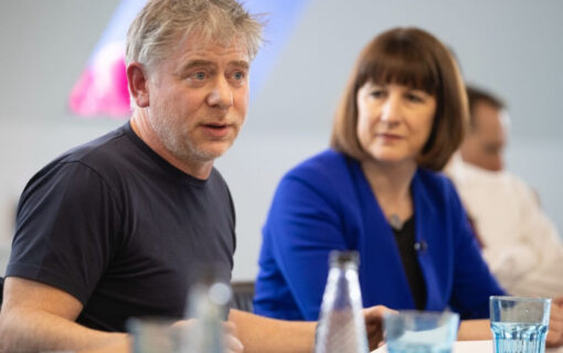 a white middle aged man in a black tshirt sits centre frame, with a woman out of focus behind him