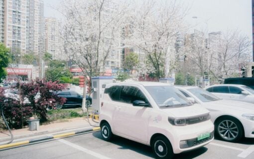 a large car plugged into an EV charger surrounded by blossoming trees and with shops with chinese lettering on in the background