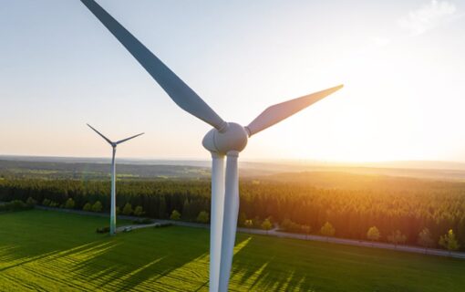 Two wind turbines, one close up and one in the distance, stood on green grass while a setting sun casts long shadows