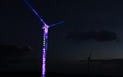 A wind turbine at night, with purple light and text reading "owned by consumers" projected onto it